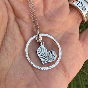 Puede incluir: Collar de plata con un colgante circular y un dije en forma de coraz&oacute;n. El dije en forma de coraz&oacute;n tiene un dise&ntilde;o de huella digital.