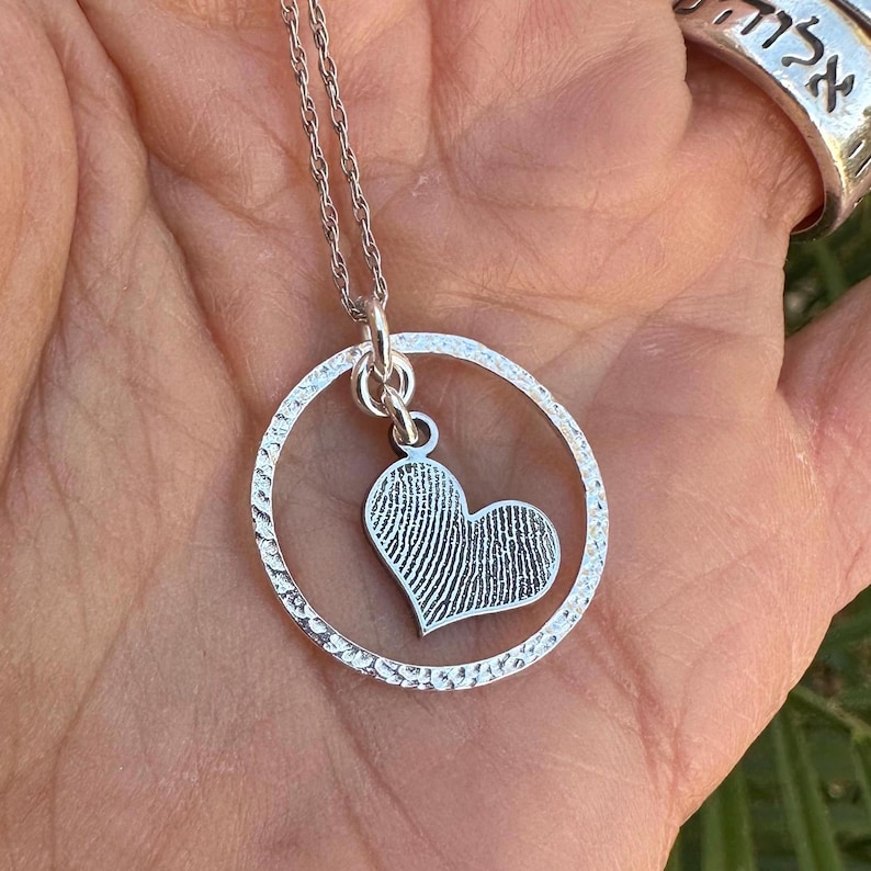 Puede incluir: Collar de plata con un colgante circular y un dije en forma de coraz&oacute;n. El dije en forma de coraz&oacute;n tiene un dise&ntilde;o de huella digital.