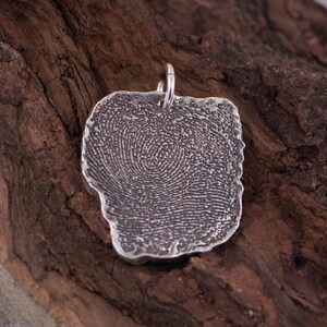 Peut inclure: Un pendentif en argent avec un motif d'empreinte digitale. Le pendentif est de forme irr&eacute;guli&egrave;re avec une surface textur&eacute;e et une petite boucle pour une cha&icirc;ne. Le motif de l'empreinte digitale est visible sur toute la surface.