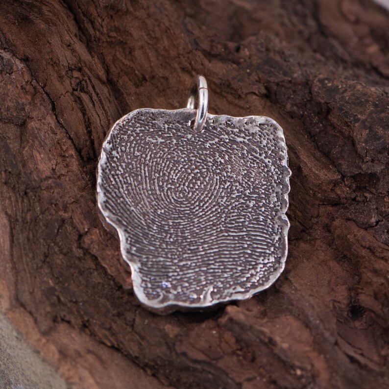 Peut inclure: Un pendentif en argent avec un motif d'empreinte digitale. Le pendentif est de forme irr&eacute;guli&egrave;re avec une surface textur&eacute;e et une petite boucle pour une cha&icirc;ne. Le motif de l'empreinte digitale est visible sur toute la surface.