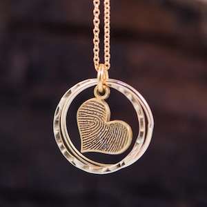 Puede incluir: Collar de oro con un colgante en forma de coraz&oacute;n con un dise&ntilde;o de huella digital. El colgante est&aacute; rodeado por dos c&iacute;rculos conc&eacute;ntricos.