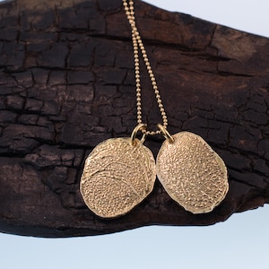 K&ouml;nnte beinhalten: Eine goldene Halskette mit zwei runden Anh&auml;ngern. Die Anh&auml;nger haben eine strukturierte Oberfl&auml;che und sind mit einer zarten Kette verbunden.