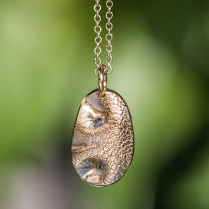 May include: A gold-toned oval pendant with a textured, dimpled surface suspended from a matching delicate link chain. The pendant features a metallic finish with subtle variations in light reflection across its uneven, organic-shaped face.