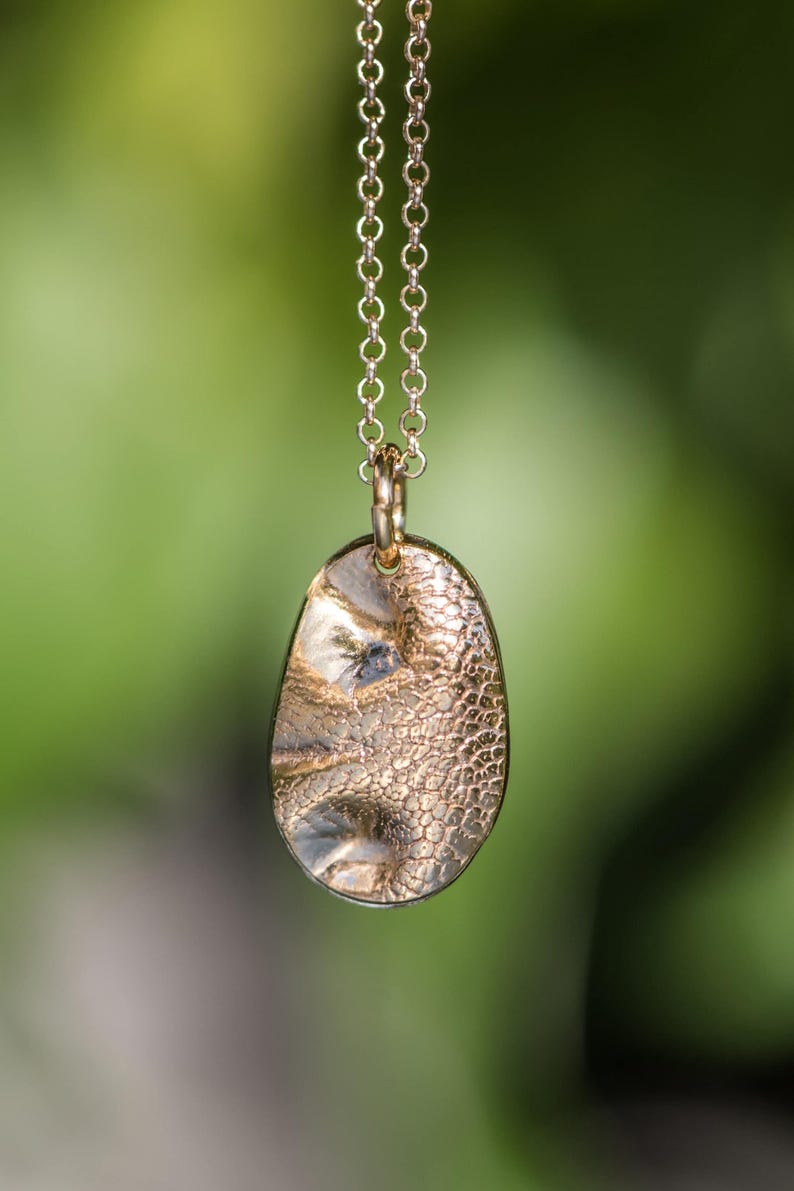 May include: A gold-toned oval pendant with a textured, dimpled surface suspended from a matching delicate link chain. The pendant features a metallic finish with subtle variations in light reflection across its uneven, organic-shaped face.