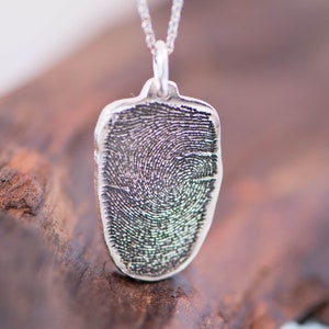 Peut inclure: Un collier avec un pendentif en argent repr&eacute;sentant une empreinte digitale. Le pendentif, de forme rectangulaire aux bords arrondis, pr&eacute;sente un motif d'empreinte digitale d&eacute;taill&eacute;. La cha&icirc;ne est &eacute;galement en argent, sur un fond en bois flou.