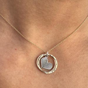 Puede incluir: Un collar de cadena dorada con un colgante en forma de coraz&oacute;n con un dise&ntilde;o de huella digital dentro de un c&iacute;rculo doble.