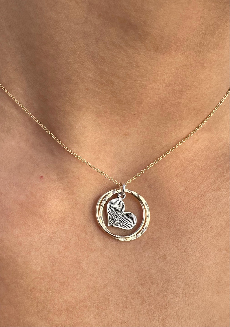 Puede incluir: Un collar de cadena dorada con un colgante en forma de coraz&oacute;n con un dise&ntilde;o de huella digital dentro de un c&iacute;rculo doble.