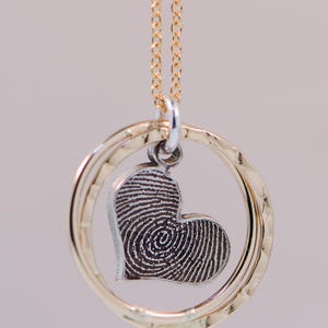 Puede incluir: Un collar de cadena dorada con un colgante de plata con un dise&ntilde;o de huella dactilar en forma de coraz&oacute;n dentro de dos anillos de oro martillados.