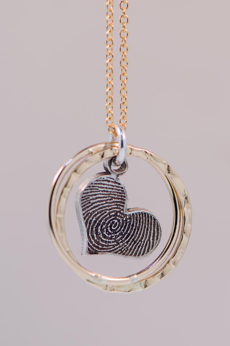 Puede incluir: Un collar de cadena dorada con un colgante de plata con un dise&ntilde;o de huella dactilar en forma de coraz&oacute;n dentro de dos anillos de oro martillados.