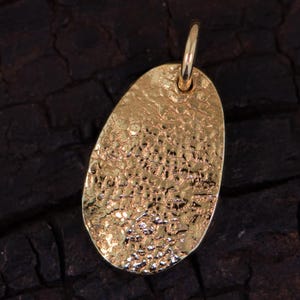 May include: A gold-toned oval pendant featuring a highly textured, hammered surface with irregular indentations and a reflective finish. The pendant includes a small, smooth circular jump ring attached at the top for hanging on a chain or cord.