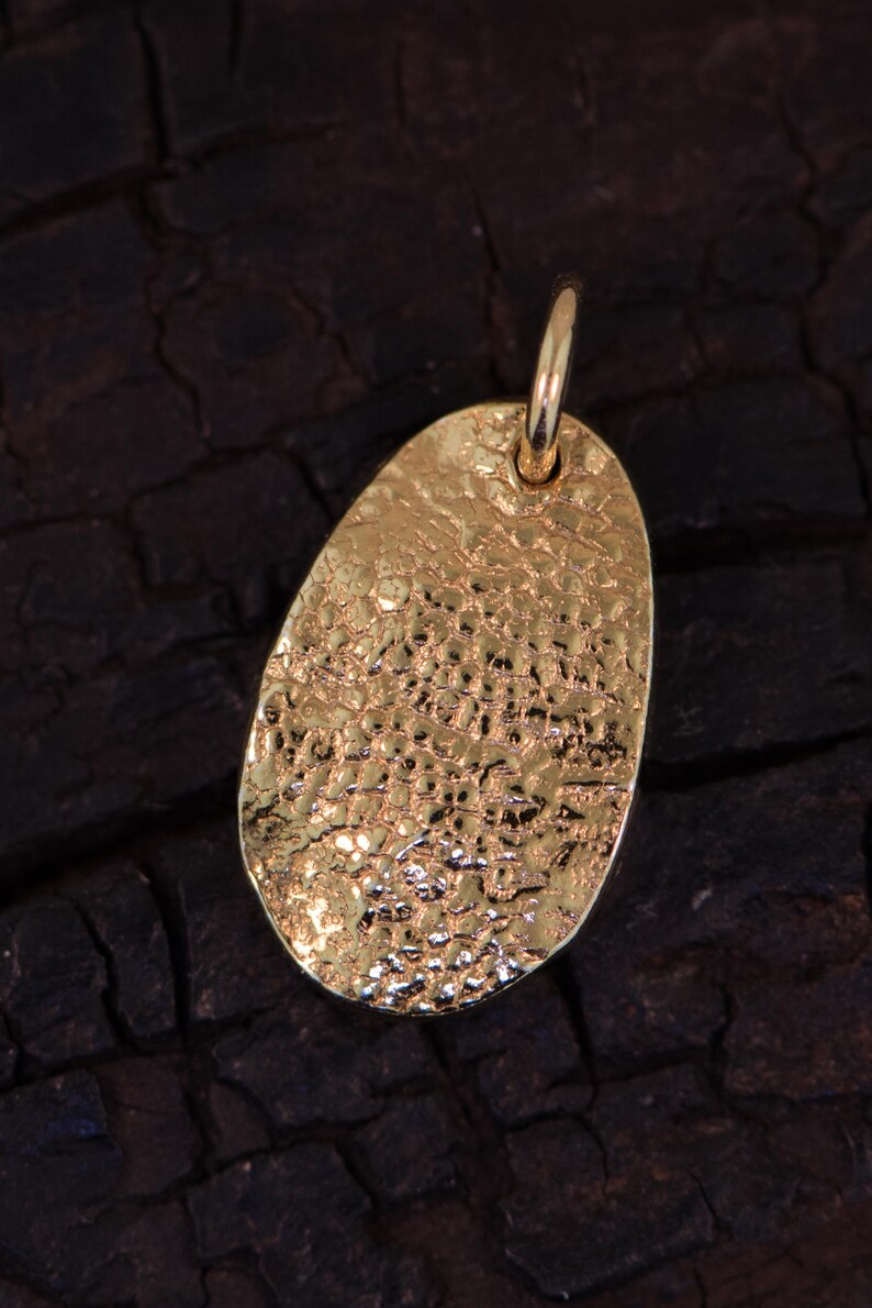 May include: A gold-toned oval pendant featuring a highly textured, hammered surface with irregular indentations and a reflective finish. The pendant includes a small, smooth circular jump ring attached at the top for hanging on a chain or cord.