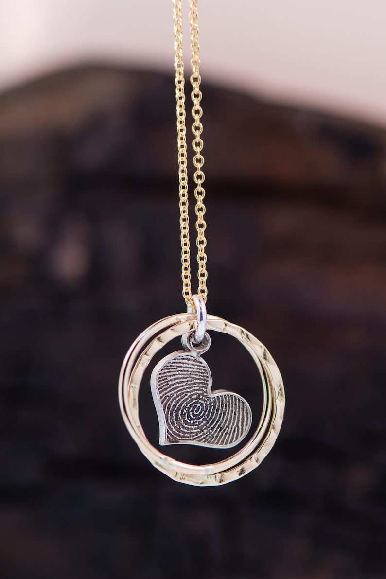 Puede incluir: Un collar de cadena dorada con un colgante de coraz&oacute;n plateado. El coraz&oacute;n tiene un dise&ntilde;o de huella digital en su interior. El colgante est&aacute; rodeado por dos anillos dorados.