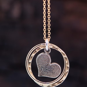 Puede incluir: Collar de cadena dorada con un colgante en forma de coraz&oacute;n con un dise&ntilde;o de huella digital. El colgante est&aacute; rodeado por dos anillos dorados.