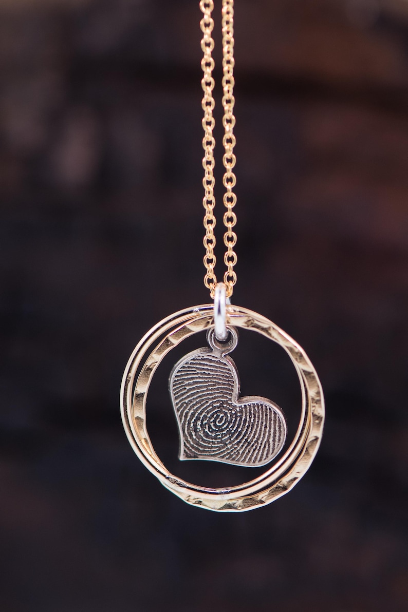 Puede incluir: Collar de cadena dorada con un colgante en forma de coraz&oacute;n con un dise&ntilde;o de huella digital. El colgante est&aacute; rodeado por dos anillos dorados.