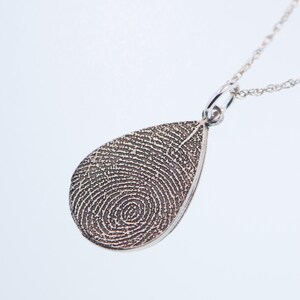 May include: Silver teardrop pendant necklace with a fingerprint design. The pendant is on a silver chain.