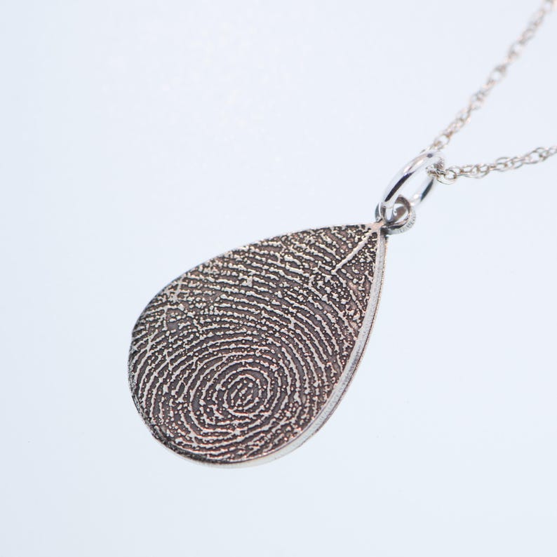 May include: Silver teardrop pendant necklace with a fingerprint design. The pendant is on a silver chain.