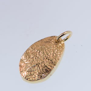 May include: A gold-toned oval pendant featuring a highly textured, hammered surface with an irregular, cratered appearance. The pendant includes a small, smooth circular jump ring attached at the top, designed for threading onto a chain or cord.