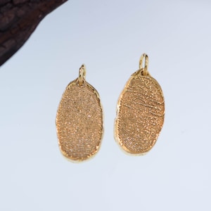 K&ouml;nnte beinhalten: Zwei ovale Anh&auml;nger in Goldfarbe mit einem Fingerabdruck-Design. Die Anh&auml;nger haben oben eine Schlaufe zum Anbringen an einer Halskette oder einem Armband.