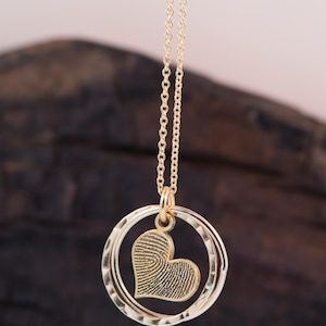 Puede incluir: Un collar de oro con un colgante en forma de coraz&oacute;n. El coraz&oacute;n est&aacute; texturizado y parece ser una huella digital. El colgante est&aacute; unido a una cadena de doble c&iacute;rculo.