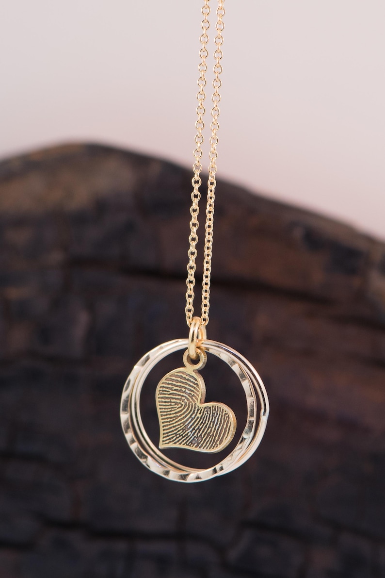 Puede incluir: Un collar de oro con un colgante en forma de coraz&oacute;n. El coraz&oacute;n est&aacute; texturizado y parece ser una huella digital. El colgante est&aacute; unido a una cadena de doble c&iacute;rculo.