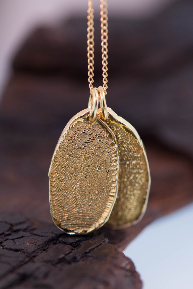 K&ouml;nnte beinhalten: Eine goldene Kettenkette mit zwei ovalen Anh&auml;ngern. Die Anh&auml;nger sind mit einem Fingerabdruckmuster strukturiert.