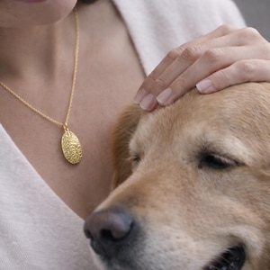 May include: A gold-toned necklace featuring a delicate chain and an oval-shaped pendant with a textured, embossed surface. The pendant hangs centrally, displaying a detailed, intricate pattern across its entire front face, suspended from a thin, polished gold-colored metal chain.