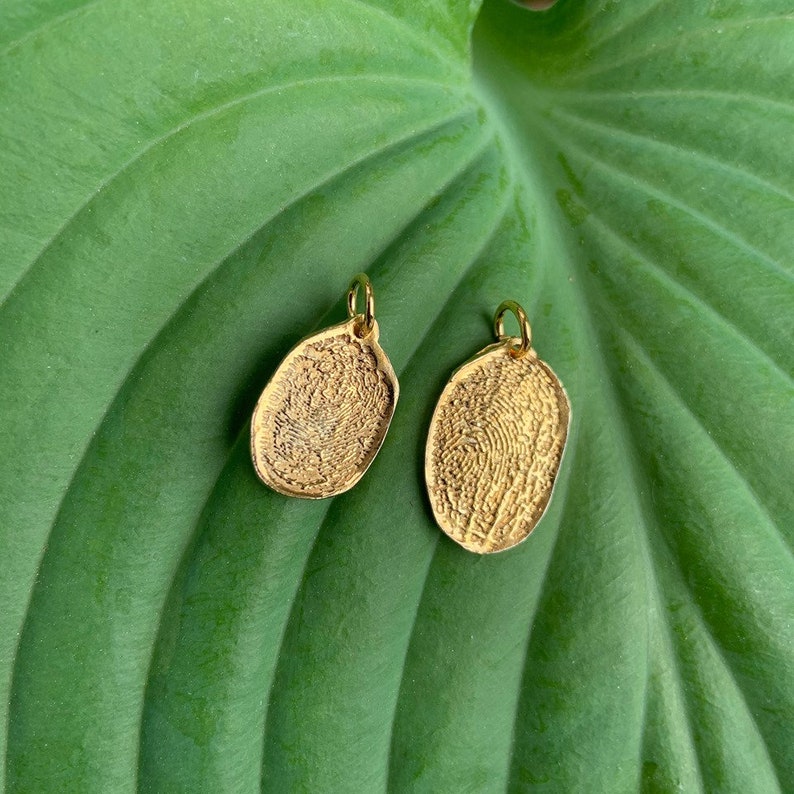 K&ouml;nnte beinhalten: Zwei goldfarbene Fingerabdruck-Anh&auml;nger, jeder mit einem einzigartigen Muster, werden auf einem gr&uuml;nen Blatt gezeigt. Die Anh&auml;nger sind oval geformt und haben eine kleine Schlaufe oben zum Anbringen an einer Halskette oder einem Armband.