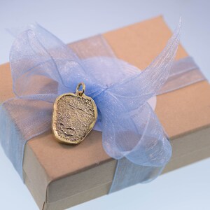 K&ouml;nnte beinhalten: Ein goldfarbener Anh&auml;nger in Herzform mit einer strukturierten Oberfl&auml;che, pr&auml;sentiert in einer braunen Geschenkbox mit einem blauen Band.