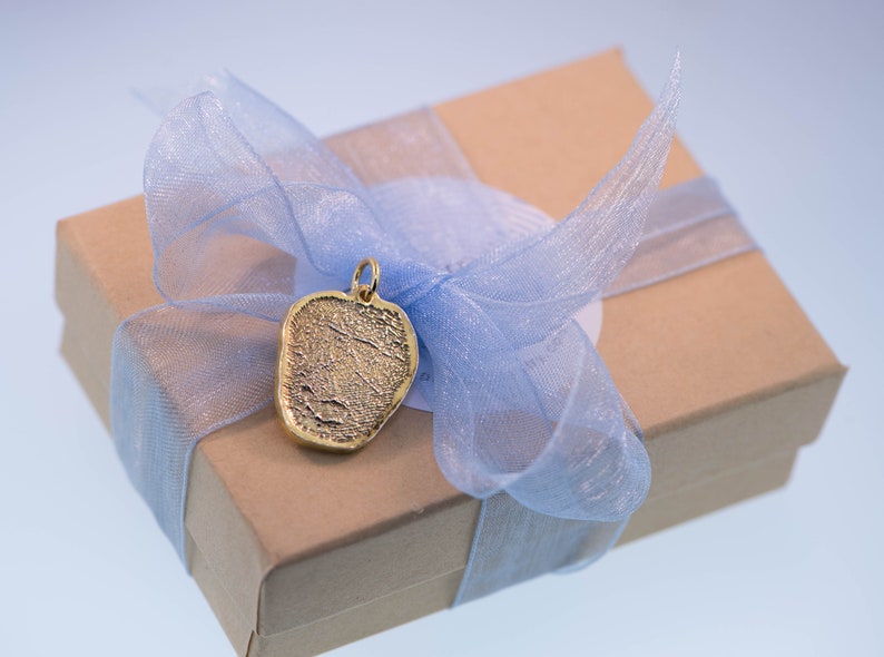 K&ouml;nnte beinhalten: Ein goldfarbener Anh&auml;nger in Herzform mit einer strukturierten Oberfl&auml;che, pr&auml;sentiert in einer braunen Geschenkbox mit einem blauen Band.