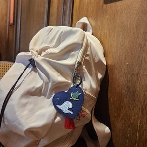 以下が含まれることがあります： ベージュの巾着バックパックと、ネイビーブルーのハート型キーホルダー。キーホルダーには、波、カメ、赤いタッセルの刺繍デザインが施されています。バックパックは木製の表面に吊り下げられています。