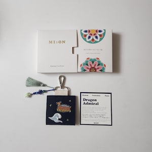 May include: A navy blue keychain with a dragon boat design, a white wave, and a tassel. The keychain is next to a white box with the text "MI:ON" and a card that says "Dragon Admiral".