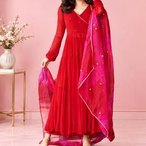 May include: A vibrant red floor-length dress with long sleeves, a V-neckline, and a tiered skirt. The dress features subtle metallic embellishments and is paired with a sheer, flowing dupatta in shades of pink and red with small mirror accents.