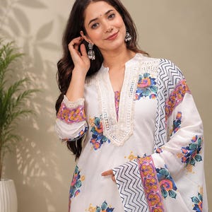 May include: A white long-sleeved tunic featuring a vibrant floral print in shades of blue, orange, and yellow. The garment includes a split neckline with cream-colored lace trim and a matching scarf with a black and white chevron border pattern.