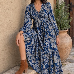 Pode incluir: Um vestido comprido de manga comprida em azul profundo com um padrão floral bege, apresentando um decote em V com acabamento em renda escura, cintura elástica e saia em camadas. O vestido é combinado com botins de camurça castanhos.