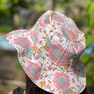 May include: A quilted bucket hat with a floral pattern. The hat has a white base with large pink flowers, green centres, and yellow details. The wide brim offers sun protection. The hat is made of a soft, quilted fabric.