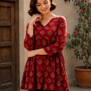 May include: A red tunic top with a V-neck and three-quarter sleeves. The top features a gold pattern and is paired with gray pants. The image is taken outdoors, with a stone wall and greenery in the background.
