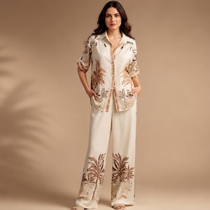 May include: A two-piece set consisting of a button-down shirt with a collar and elbow-length sleeves, paired with wide-leg trousers. Both pieces are cream-coloured and feature a repeating brown palm tree and tropical foliage print pattern.