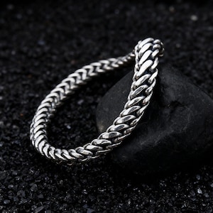 May include: A silver-colored chain bracelet with a woven design. The bracelet is resting on a black stone against a dark background. The chain links are closely woven together, creating a textured appearance.