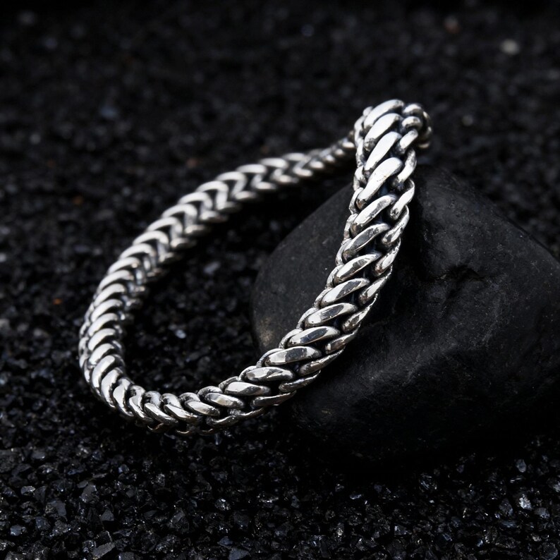 May include: A silver-colored chain bracelet with a woven design. The bracelet is resting on a black stone against a dark background. The chain links are closely woven together, creating a textured appearance.