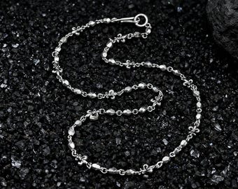 May include: A silver necklace with a delicate chain design. The necklace features small, faceted beads and fleur-de-lis charms. The clasp is a simple hook and eye closure. The jewelry is displayed on a dark, textured surface.
