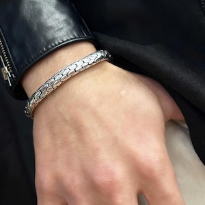 May include: A silver bracelet with a scale-like texture. The bracelet is worn on a wrist, with a black leather jacket in the background. The bracelet appears to be made of metal.