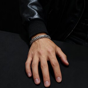 May include: A silver-colored, chain-link bracelet is worn on a person's wrist. The bracelet is made of multiple, thick, interlocking links. The person is wearing a black leather jacket and a black sweater.