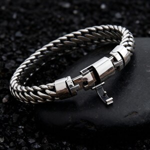 May include: A silver-colored bracelet with a woven chain design. The bracelet features a secure clasp mechanism. The jewelry is displayed on a dark, textured surface, highlighting the metallic sheen of the bracelet.