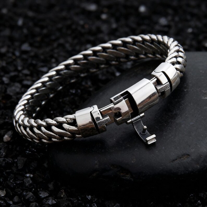 May include: A silver-colored bracelet with a woven chain design. The bracelet features a secure clasp mechanism. The jewelry is displayed on a dark, textured surface, highlighting the metallic sheen of the bracelet.