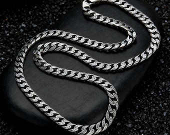 May include: A silver-colored chain necklace with a detailed, textured pattern. The necklace is displayed on a dark, textured surface, showcasing its intricate design and metallic sheen. The chain is a classic jewelry piece.