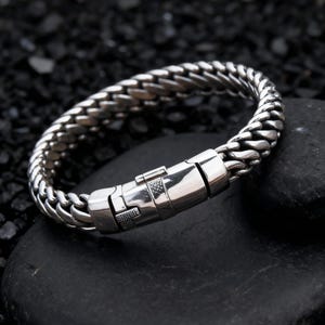 May include: A silver-colored bracelet with a woven chain design. The bracelet features a secure clasp closure. The jewelry is displayed on a black, textured surface.