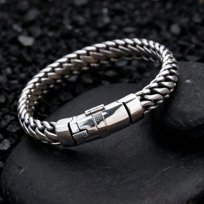 May include: A silver-colored bracelet with a woven chain design. The bracelet features a secure clasp closure. The jewelry is displayed on a black, textured surface.