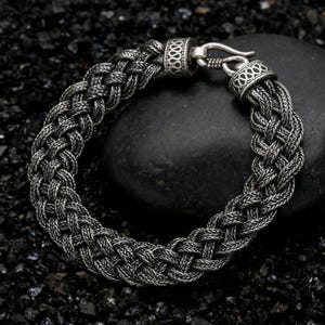 Puede incluir: Una pulsera trenzada plateada con cierre de gancho. La pulsera presenta un diseño grueso y tejido con detalles intrincados. El cierre y los extremos tienen patrones decorativos. La pulsera se muestra sobre una superficie oscura y texturizada.