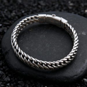 May include: A silver-colored bracelet with a curb chain design. The bracelet is made of metal and features a secure clasp. The bracelet is displayed on a black stone surface.