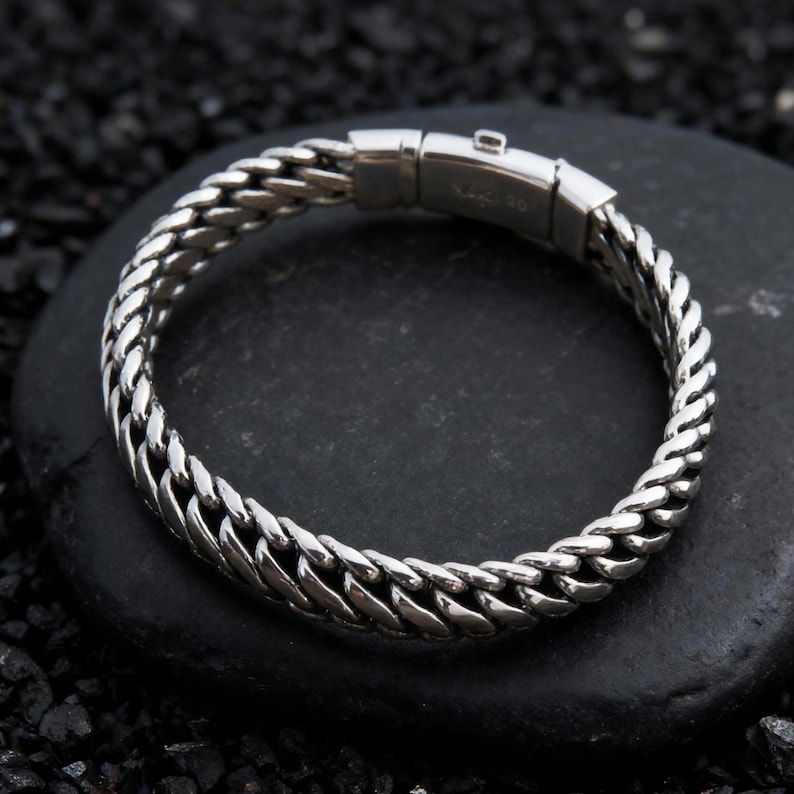 May include: A silver-colored bracelet with a curb chain design. The bracelet is made of metal and features a secure clasp. The bracelet is displayed on a black stone surface.